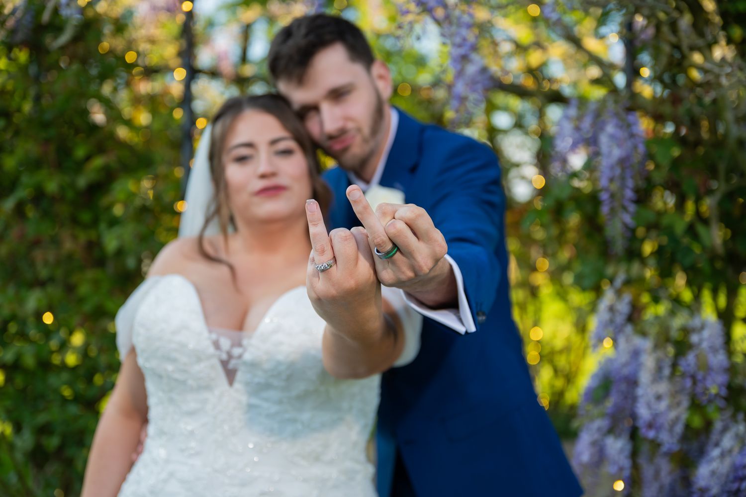 Bride and groom showing off wedding rings outdoors under wisteria blooms in a romantic garden setting.