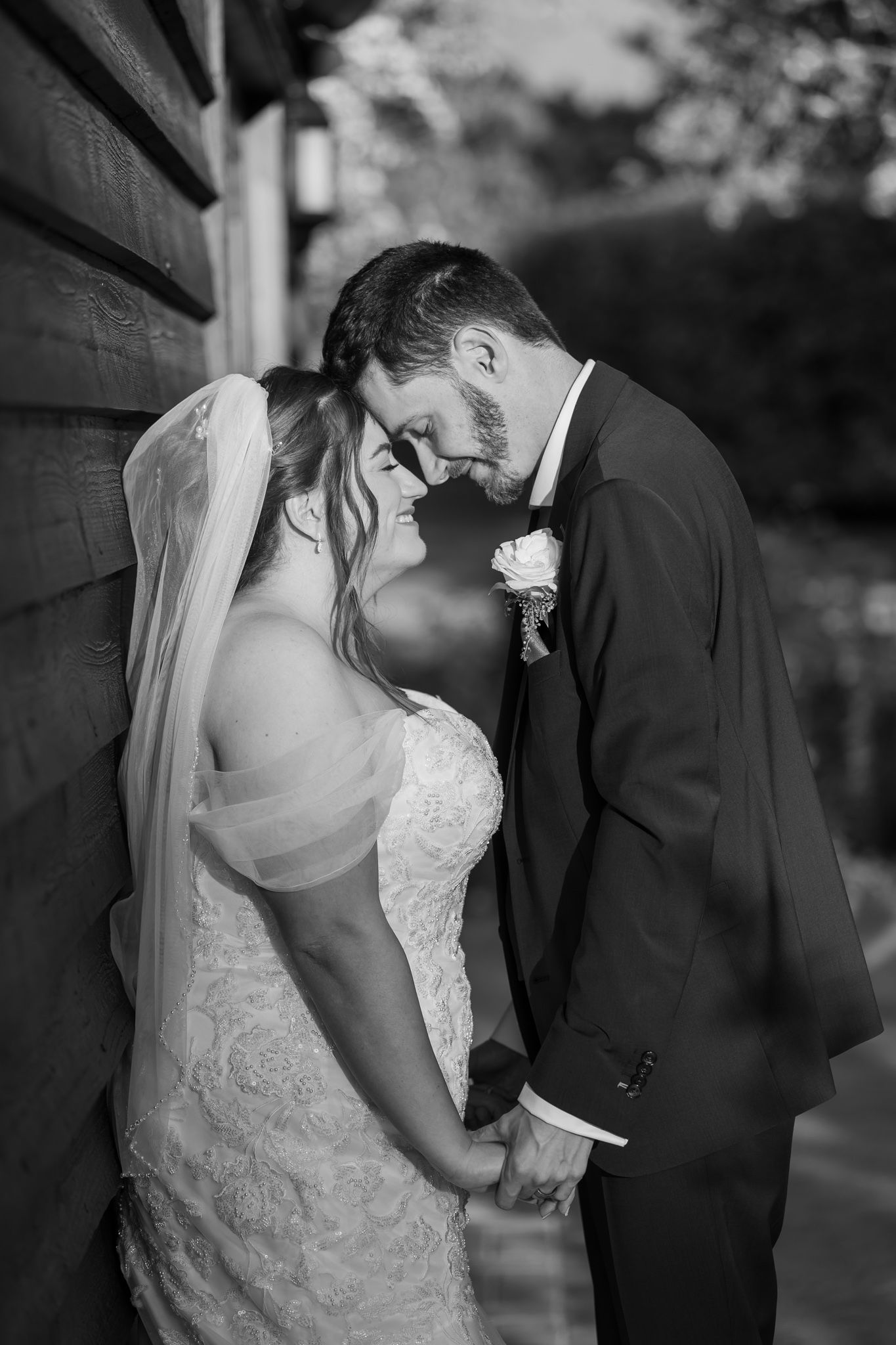 Black and white wedding photo of bride and groom holding hands, foreheads touching tenderly against a rustic wooden wall.
