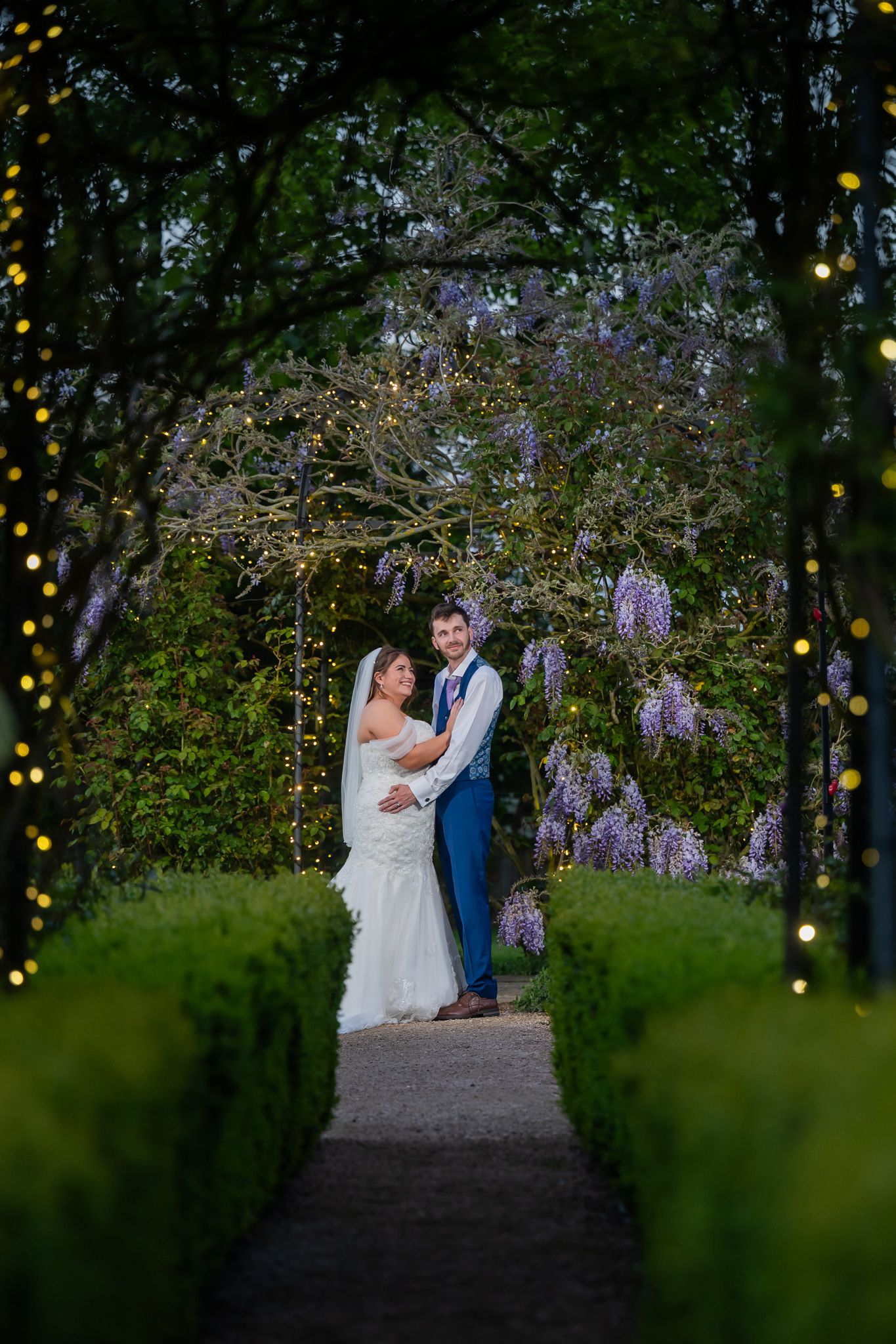 Bride and groom embrace under a wisteria arch with fairy lights in a lush garden at dusk.