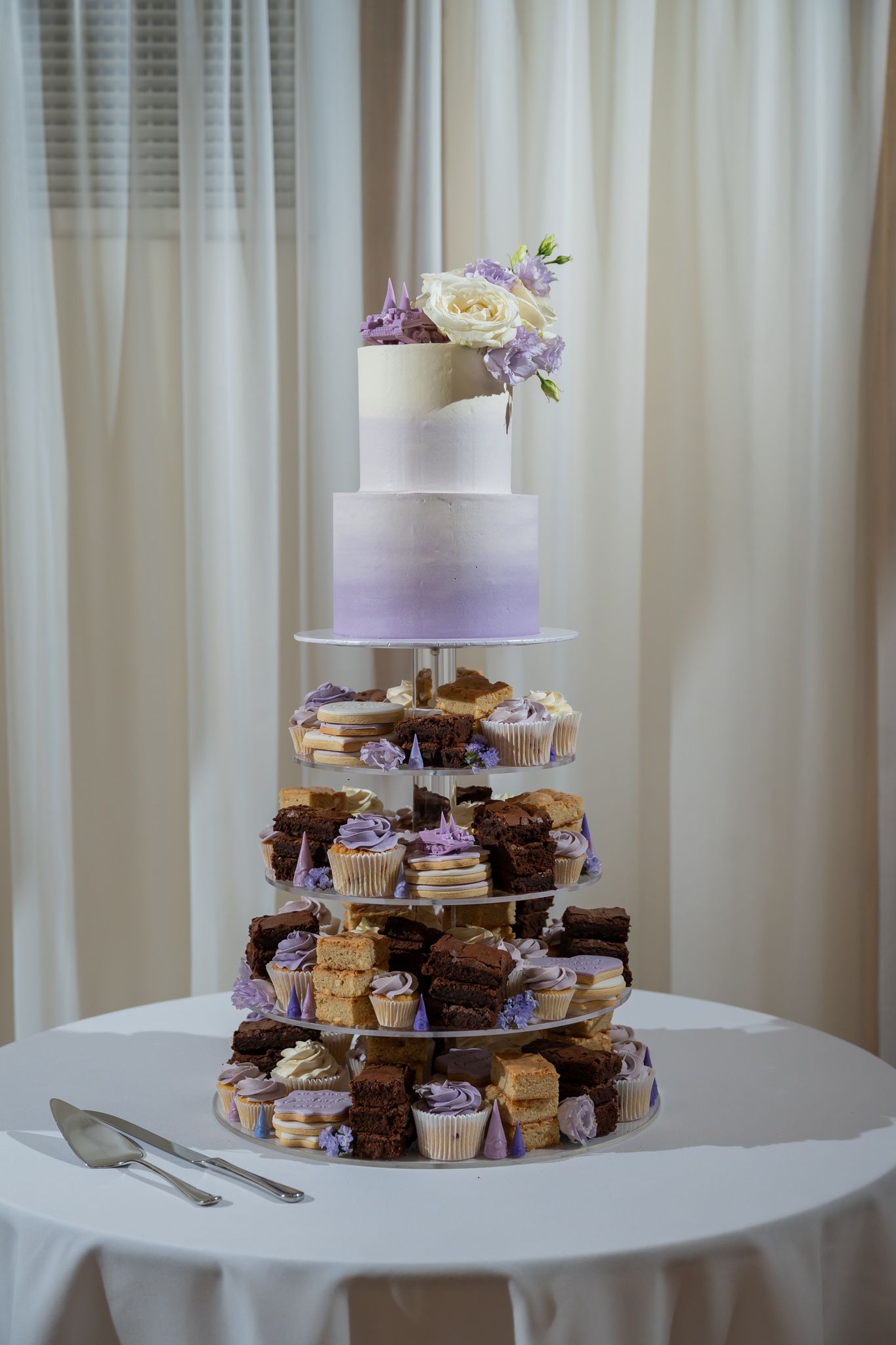 Elegant wedding cake tower with purple ombre tiers, cupcakes, and brownies topped with white roses on a draped table.