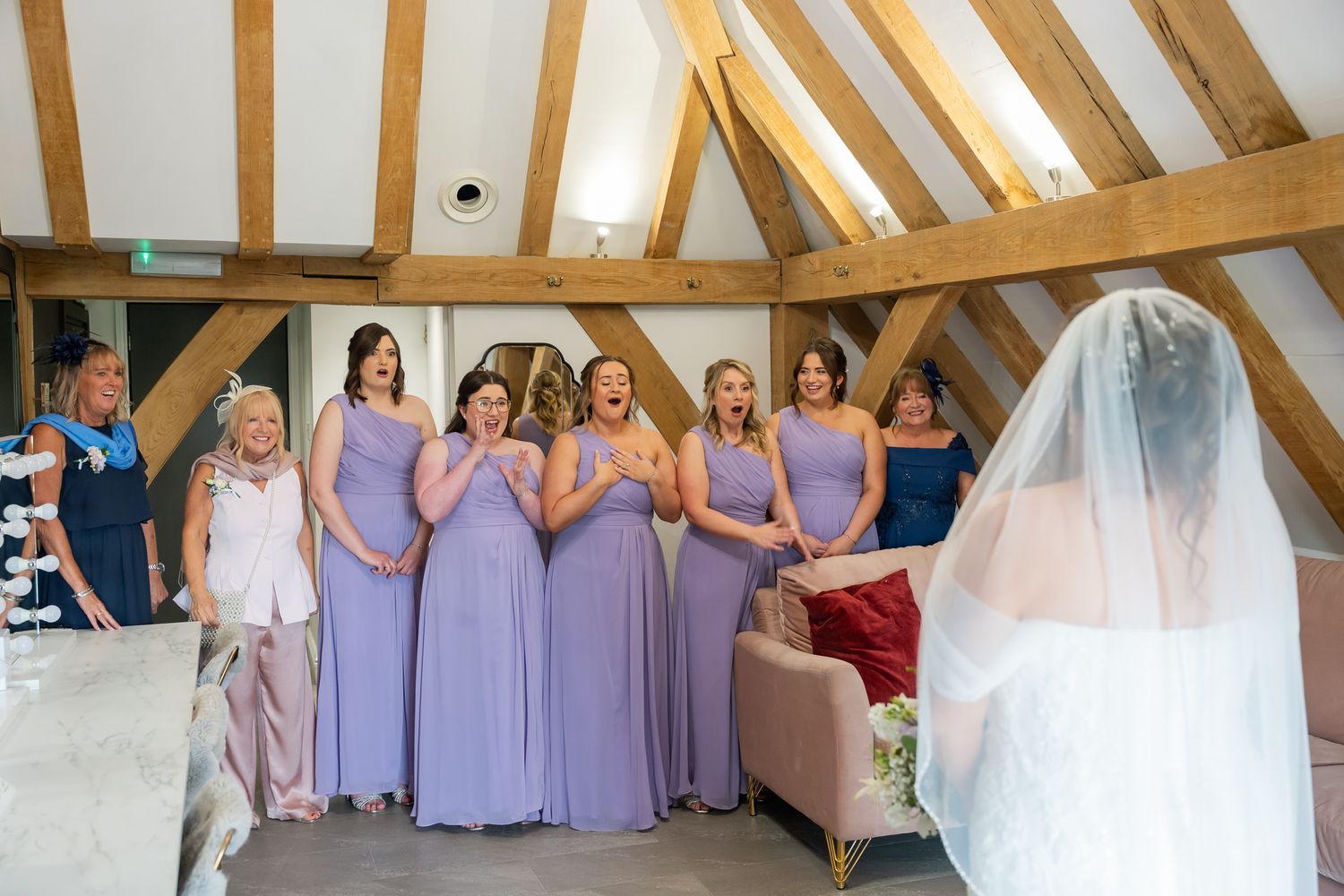 Bridesmaids in lavender gowns react with joy and surprise as bride reveals her wedding dress in the Bridal suite at Old Kent Barn