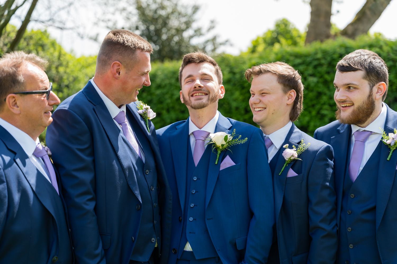 Groom and groomsmen in navy blue suits with lavender ties and white boutonnieres laughing outdoors.