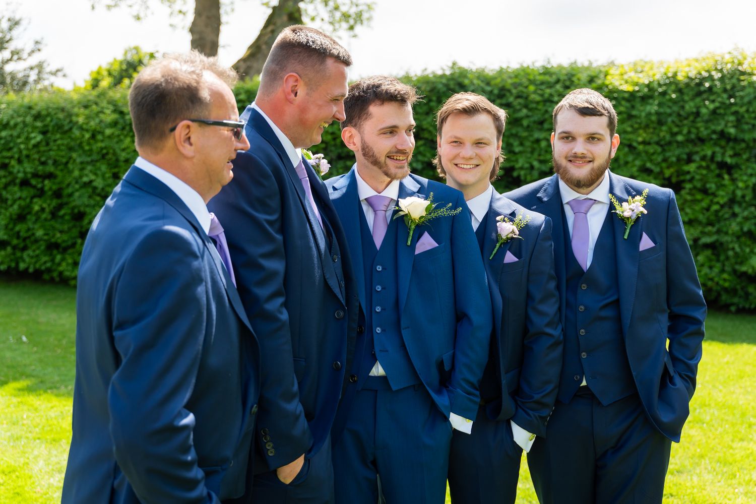 Five groomsmen in navy blue suits with purple boutonnieres laughing together outdoors at a wedding at Old Kent Barn in Kent.