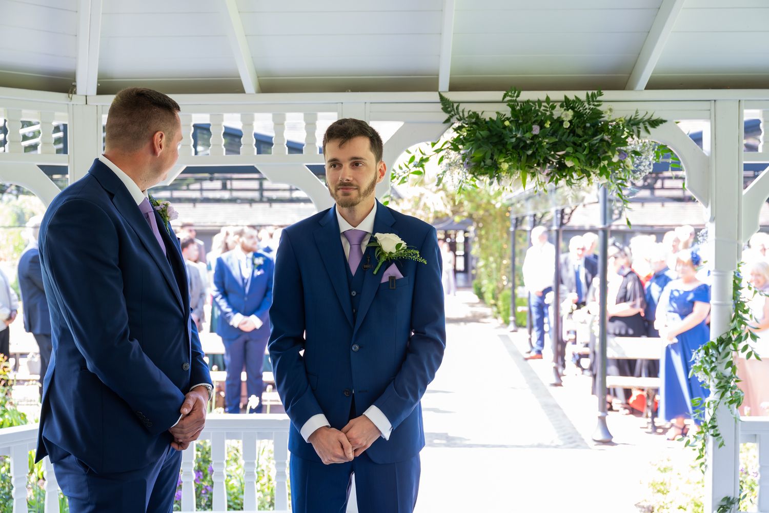 Groom in navy blue suit with boutonniere awaits bride at outdoor wedding ceremony gazebo with guests seated behind.