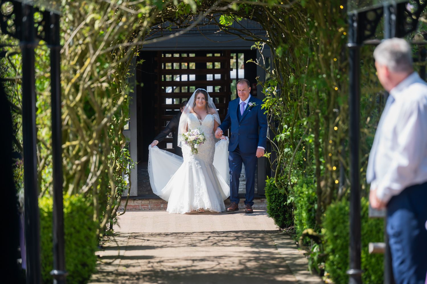 Bride and her father walking through the floral archway for the wedding in the gardens at Old Kent Barn in Kent.