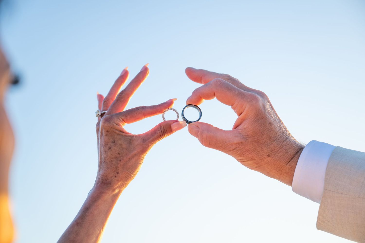 Two hands exchanging wedding rings against a clear blue sky, symbolizing a marriage ceremony.