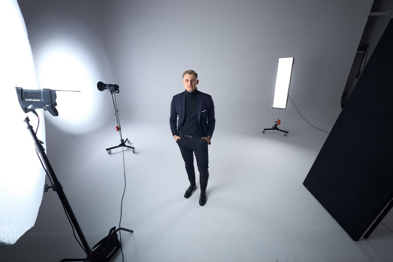 Man in dark suit stands confidently in professional photo studio surrounded by lighting equipment on white backdrop.