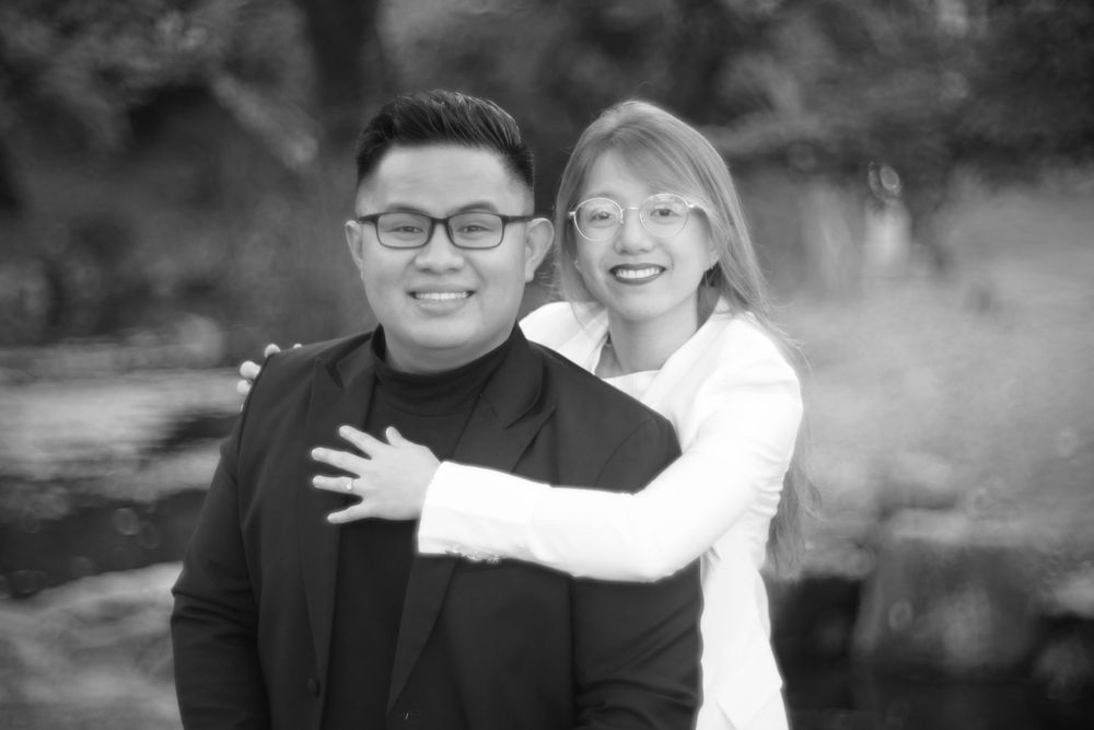 Black and white photo of a smiling couple posing together outdoors, woman hugging man from behind.