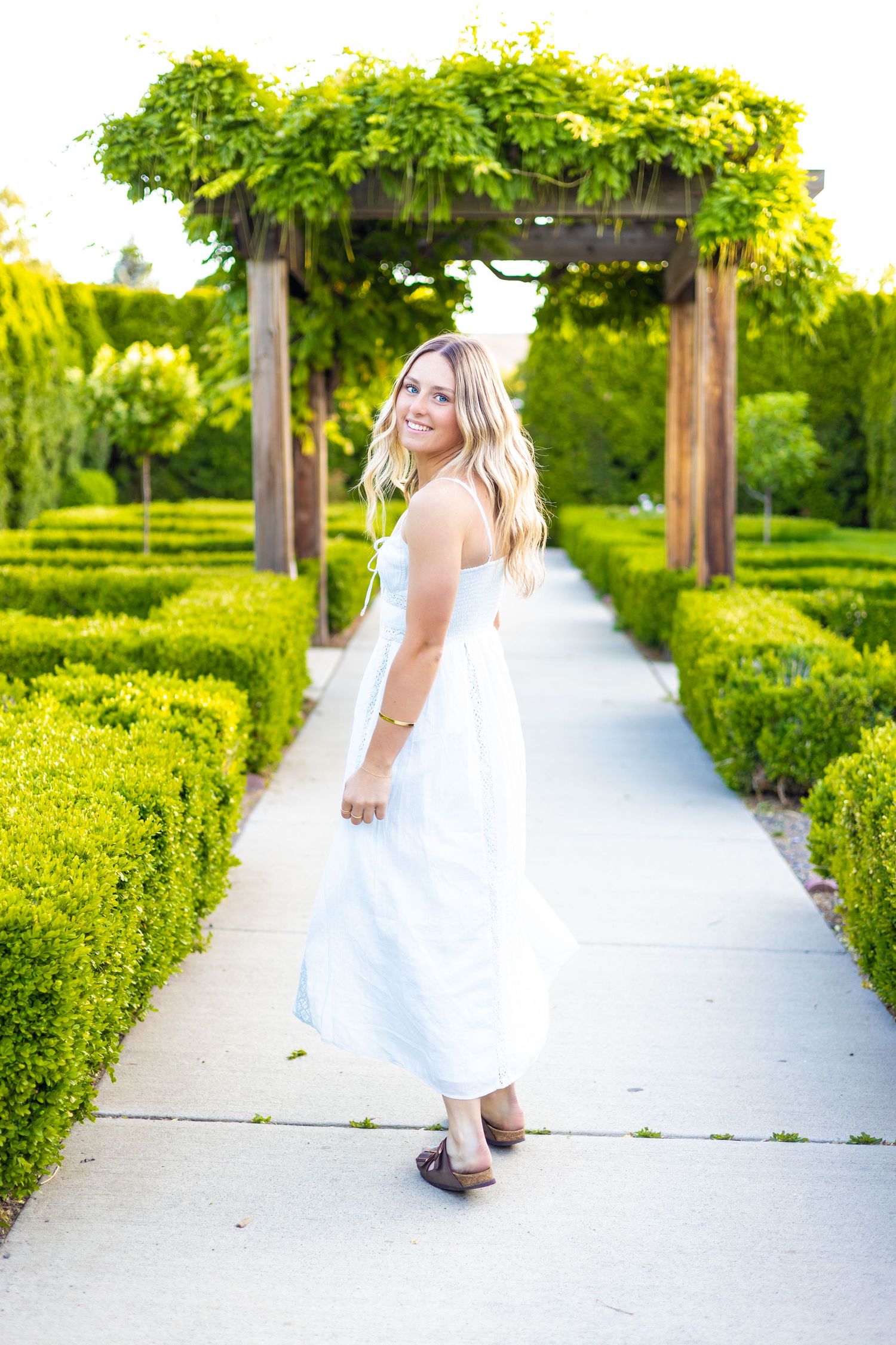 Smiling blonde woman in white maxi dress posing on garden pathway with green hedges and wooden pergola.