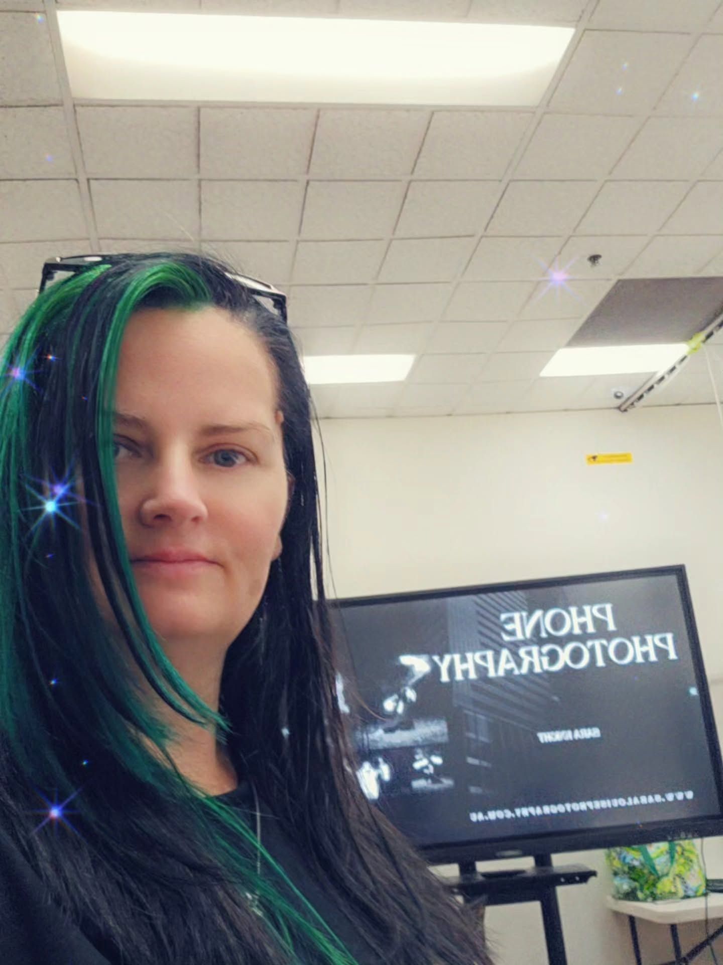 Woman with black and green dreadlocks takes selfie in front of Phone Photography presentation screen in classroom.