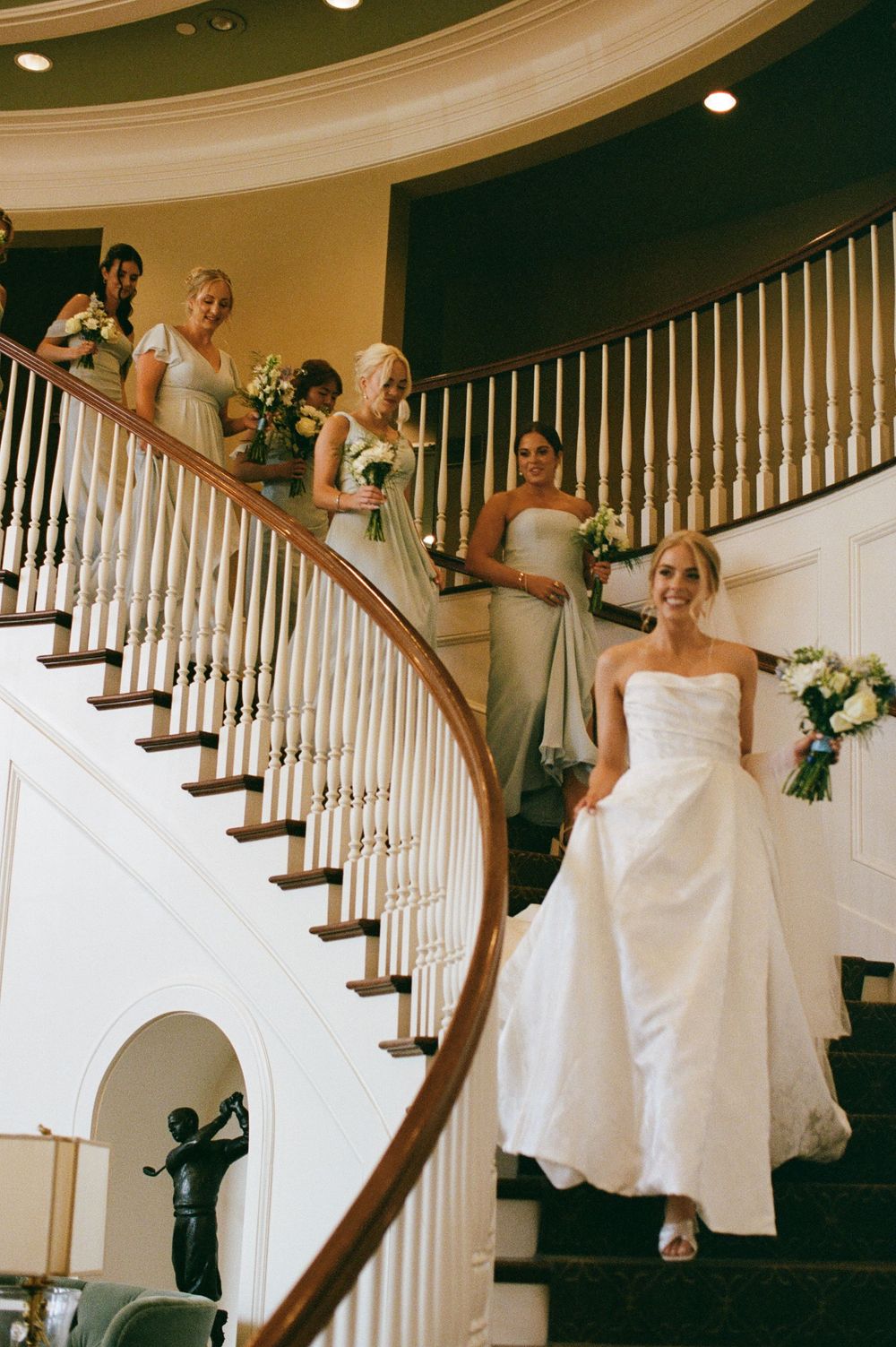 Bride in white strapless gown descends elegant curved staircase with bridesmaids in sage green dresses holding bouquets.