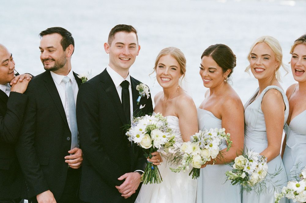 Bride and groom with wedding party smiling by the water, bridesmaids holding white floral bouquets in blue dresses.