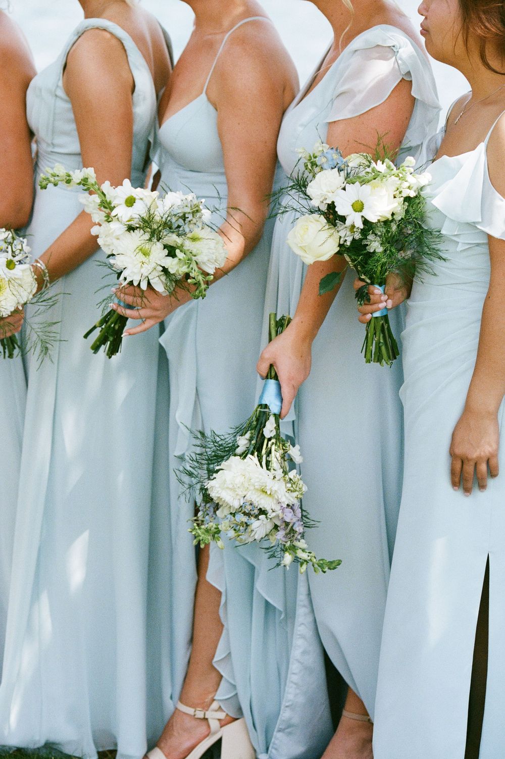Close-up of bridesmaids in pale blue chiffon gowns holding white floral bouquets with greenery accents.