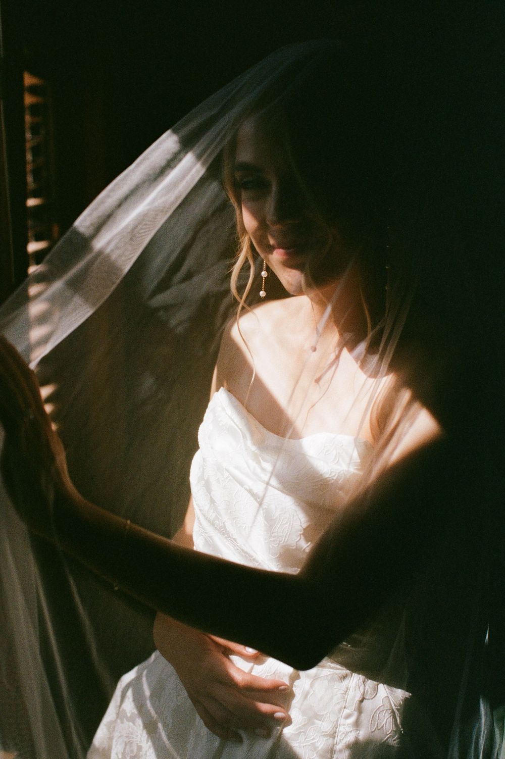 A bride in a white dress and veil stands in soft sunlight, wearing delicate drop earrings and a necklace.