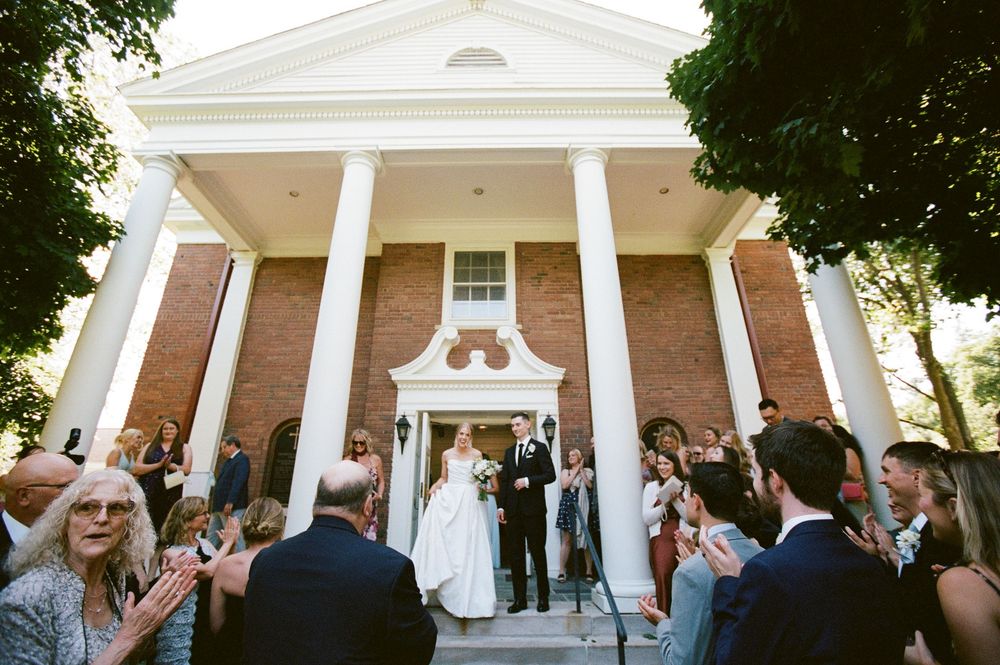 Newlyweds exit brick colonial church with white columns as guests celebrate outside after ceremony.