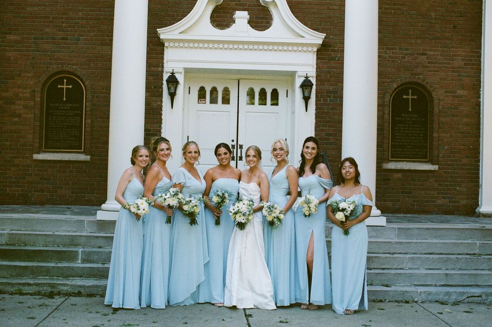 Bride in white gown poses with six bridesmaids in light blue dresses outside a brick church.