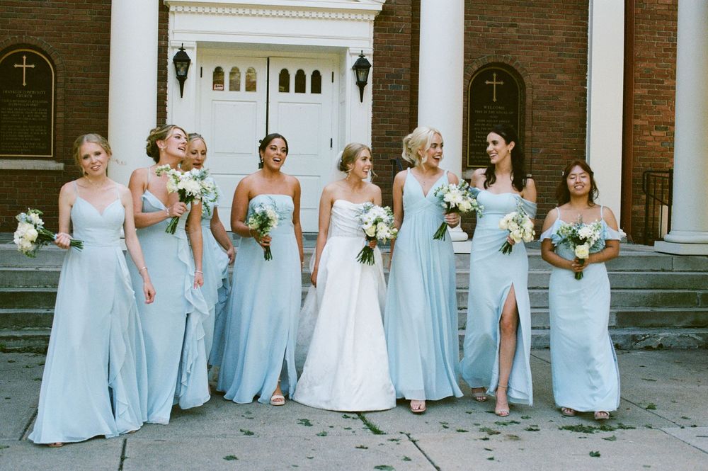 Bride and bridesmaids in light blue gowns laugh together outside a white-doored brick church.