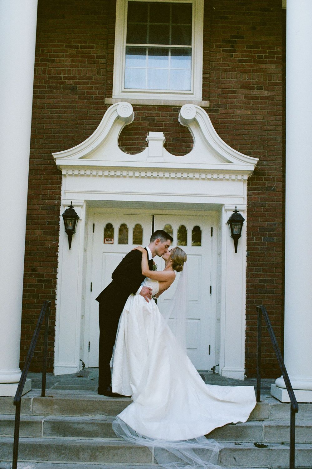Bride and groom share a kiss on steps of brick church with white colonial doorway.