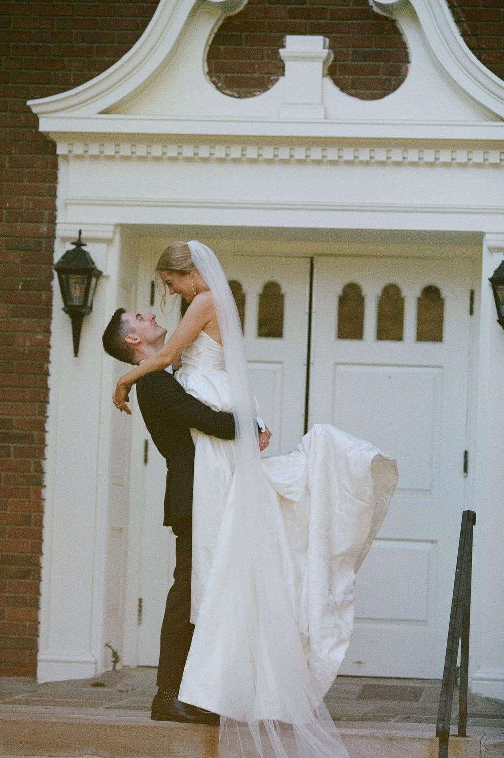 Groom lifting bride in white wedding gown and veil outside elegant church doors.