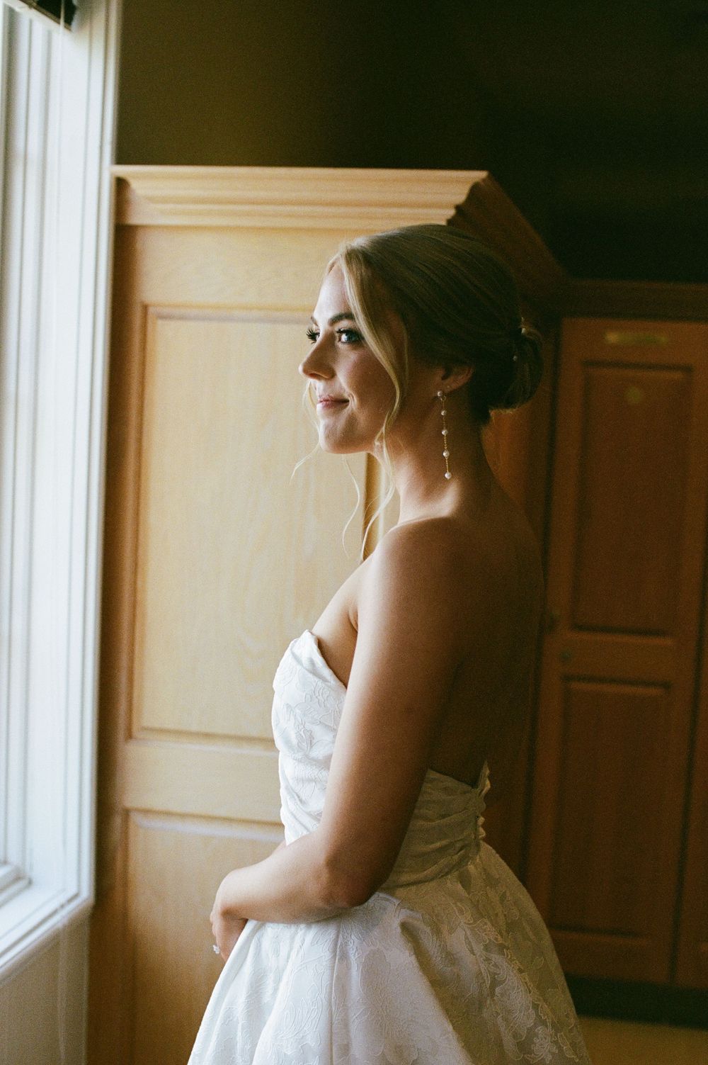 Elegant bride in strapless lace gown with updo and drop earrings gazes out window in soft natural light.