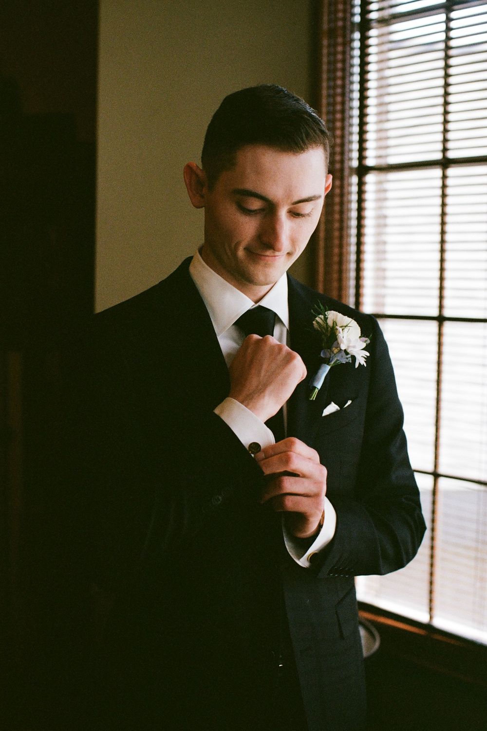 Dapper groom in black suit adjusts boutonniere with white flower near window with soft natural lighting.