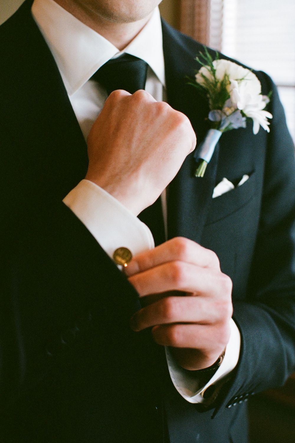 Groom adjusts cufflinks on white shirt with boutonniere on black suit.