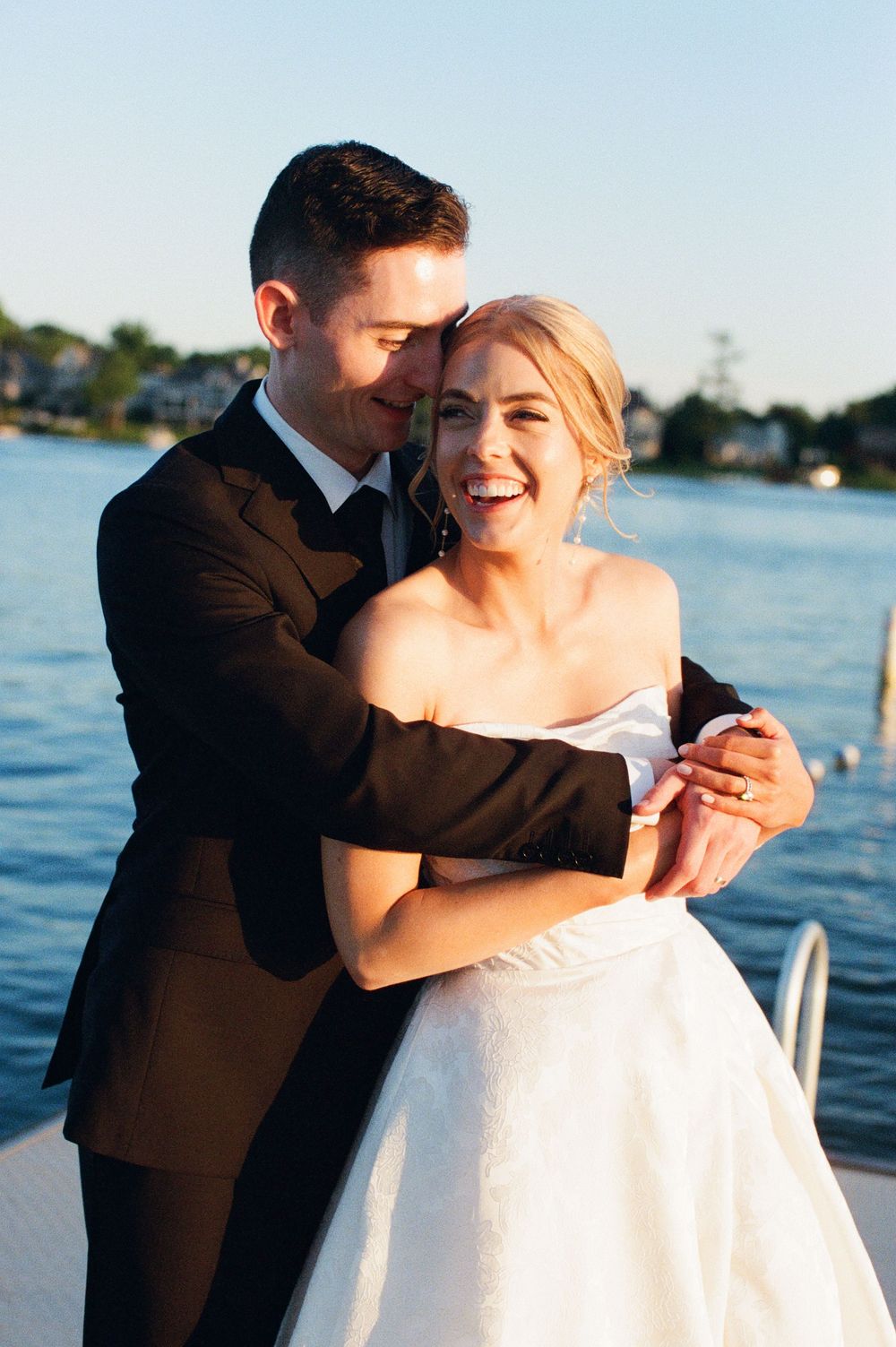 A joyful bride in a white strapless gown embraces her groom in a dark suit on a waterfront dock at golden hour.