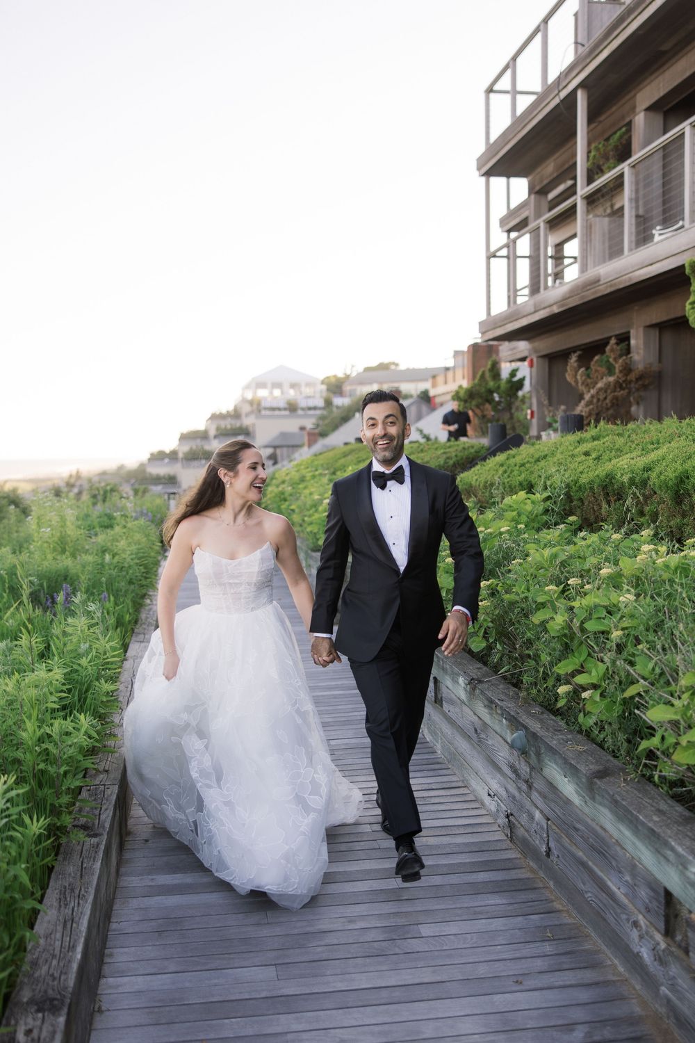 Bride and groom walk hand-in-hand on a wooden boardwalk surrounded by lush greenery near a coastal venue.