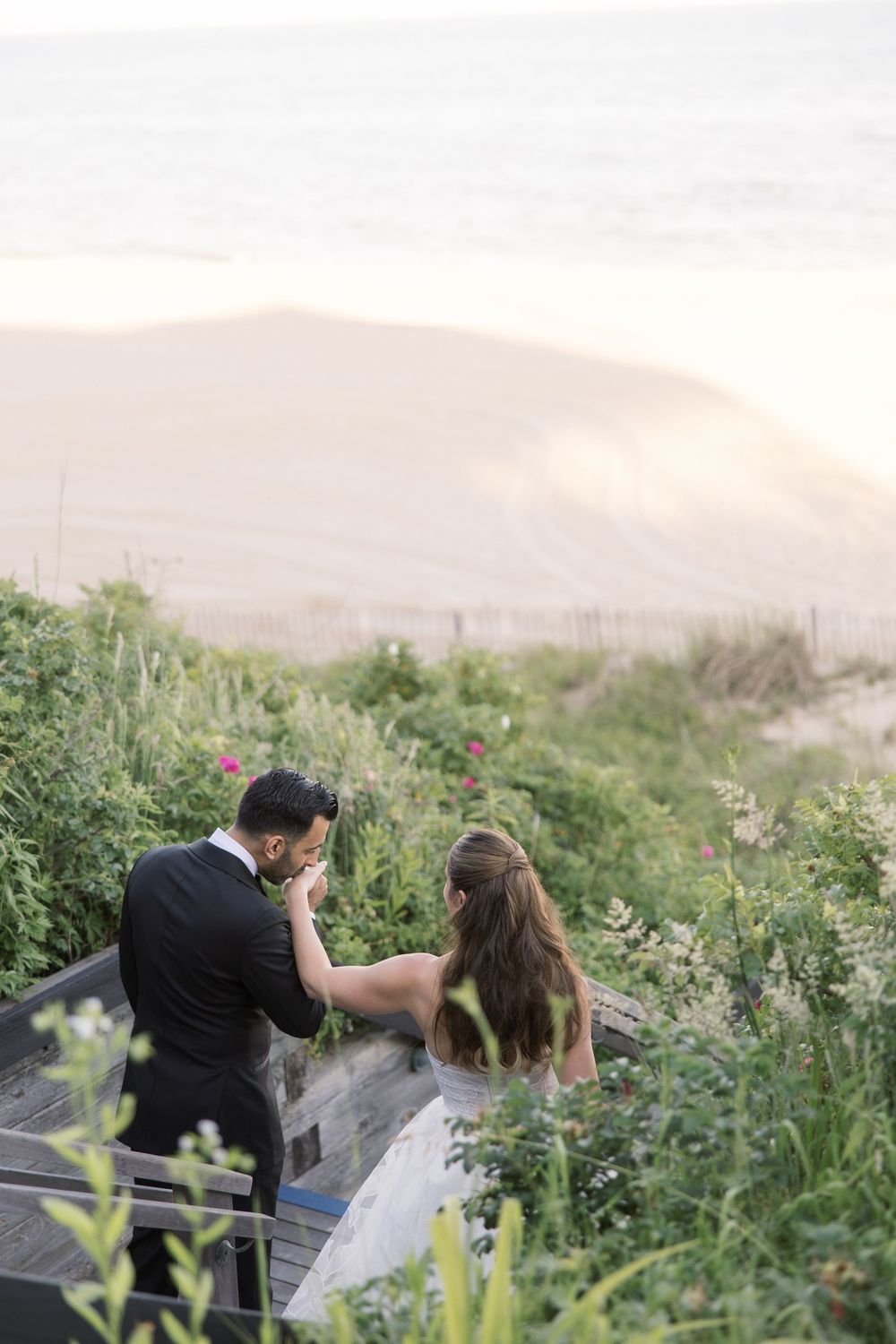 Bride and groom walk hand-in-hand down beach stairs surrounded by lush greenery with sandy dunes and ocean in background.