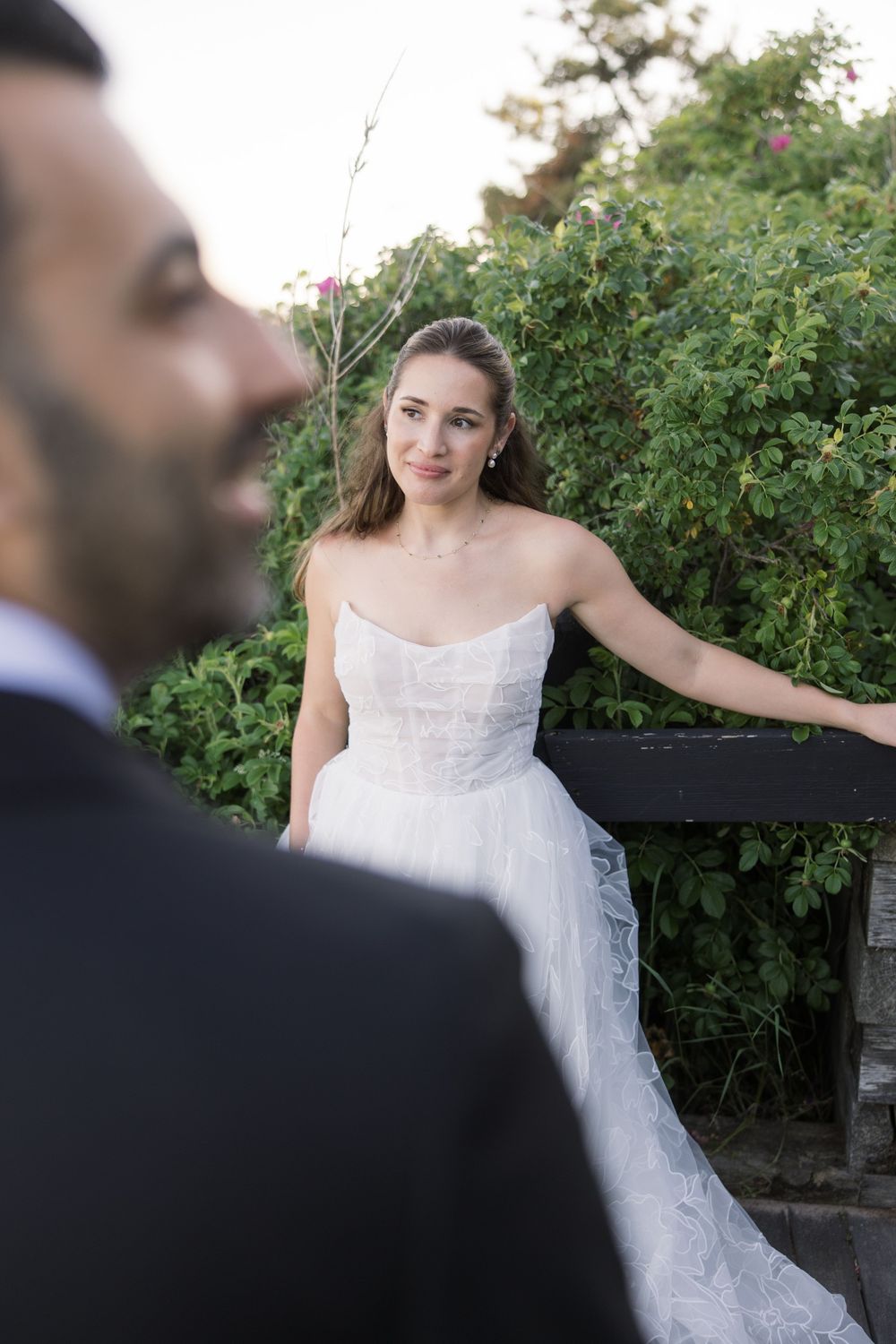 Bride in strapless white gown leans on fence surrounded by greenery, groom in foreground at outdoor wedding.