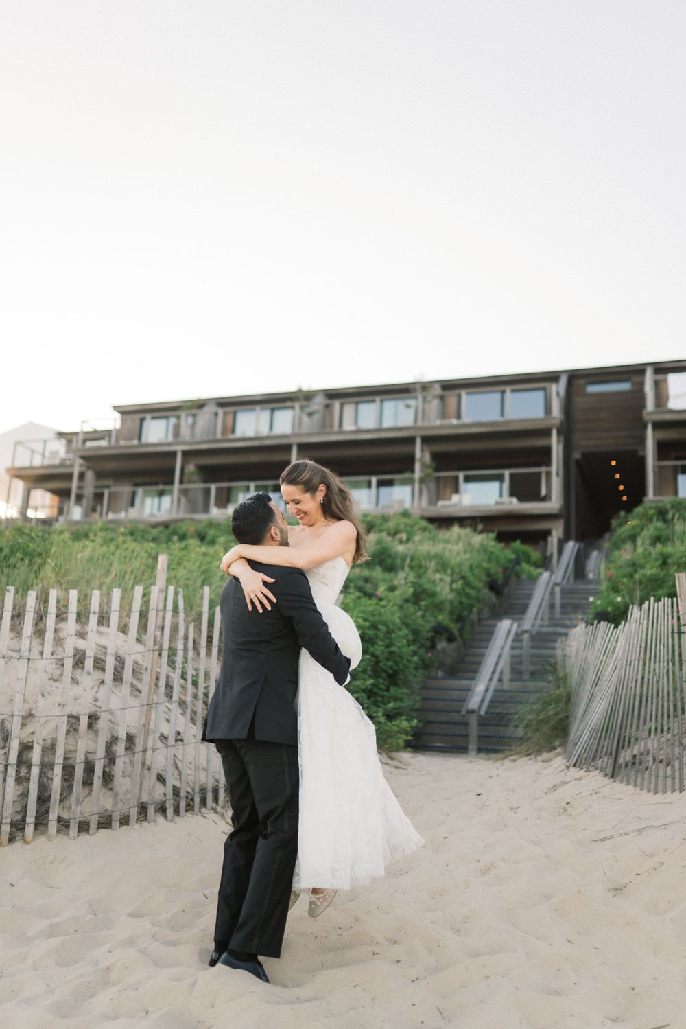 Bride and groom embrace on sandy beach with coastal wedding venue in background at sunset.