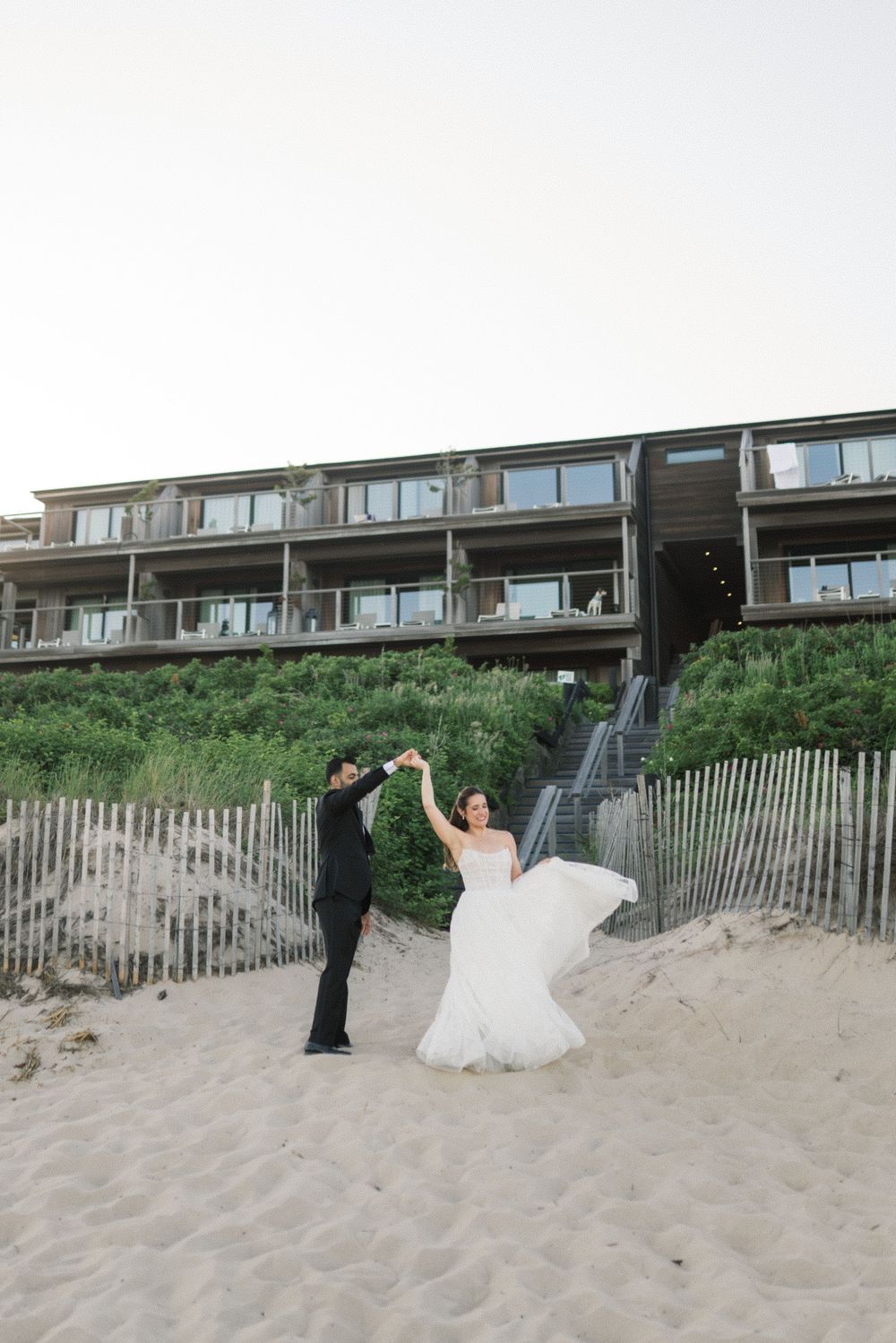 Bride and groom dancing on sandy beach in front of coastal hotel, wedding dress flowing in the wind.