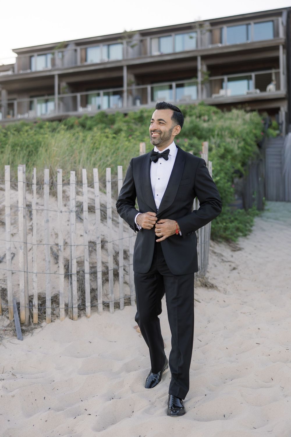 Groom in black tuxedo and bow tie smiling on sandy beach with wooden fence and building in background.