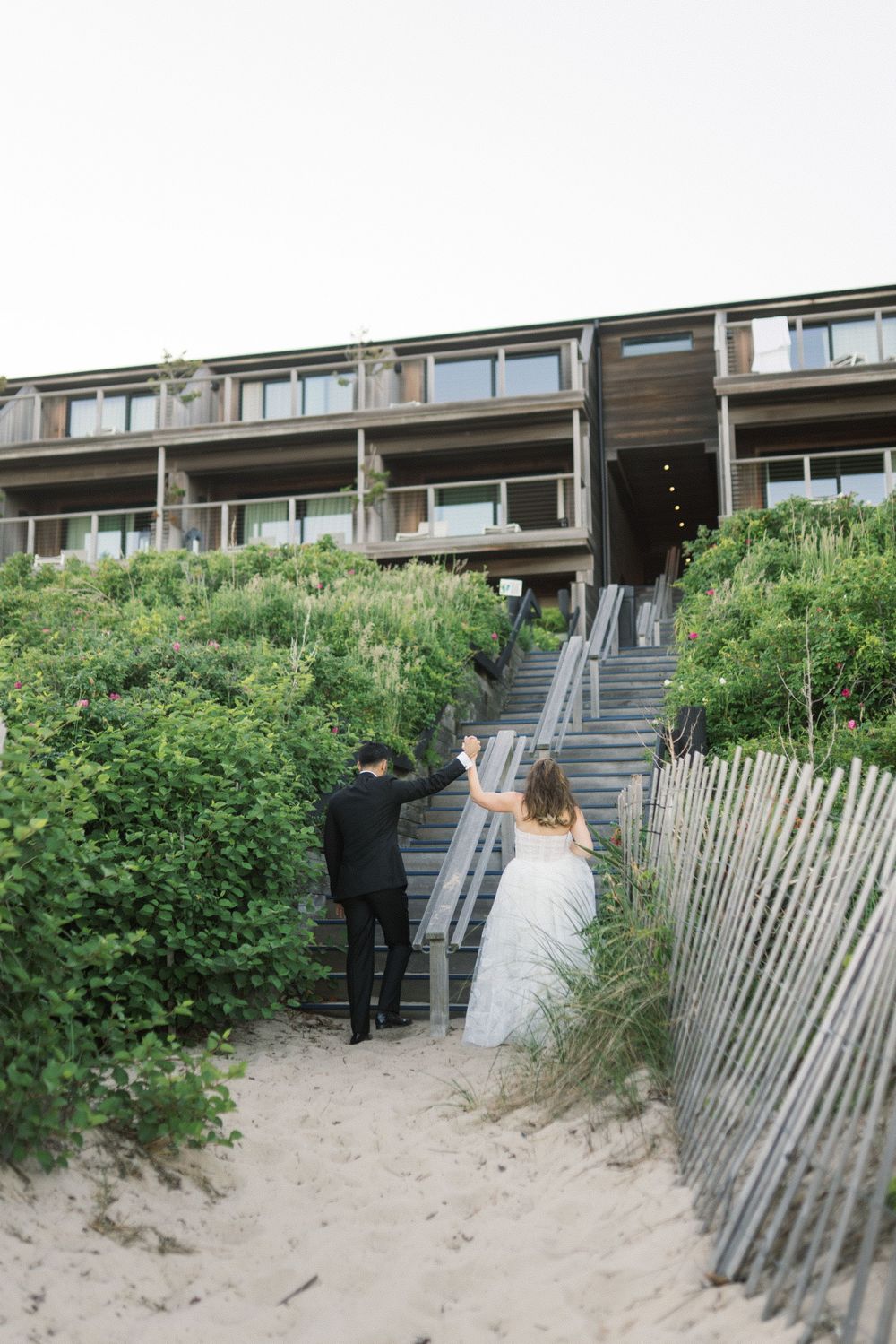 Bride and groom hold hands walking toward beachfront hotel stairway lined with dune fencing and green shrubs.