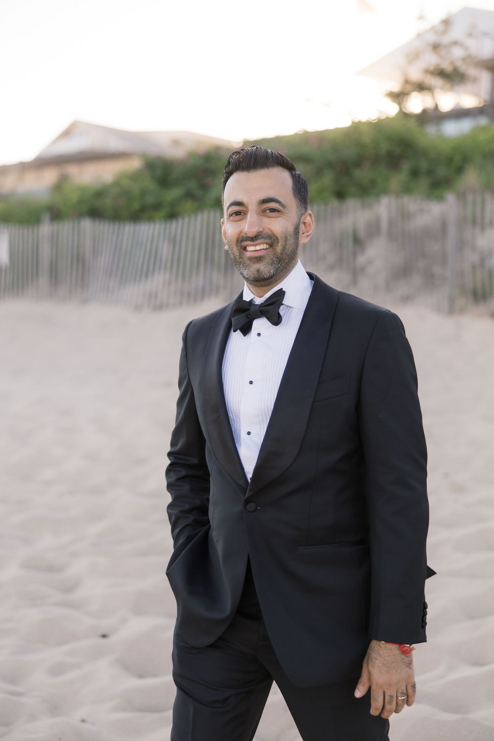 Smiling groom in black tuxedo with bow tie standing on sandy beach at sunset.