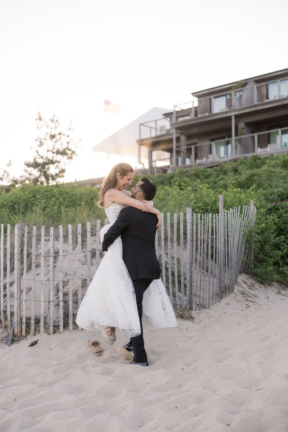 Groom lifting bride in white wedding dress on sandy beach with wooden fence and coastal house in background.