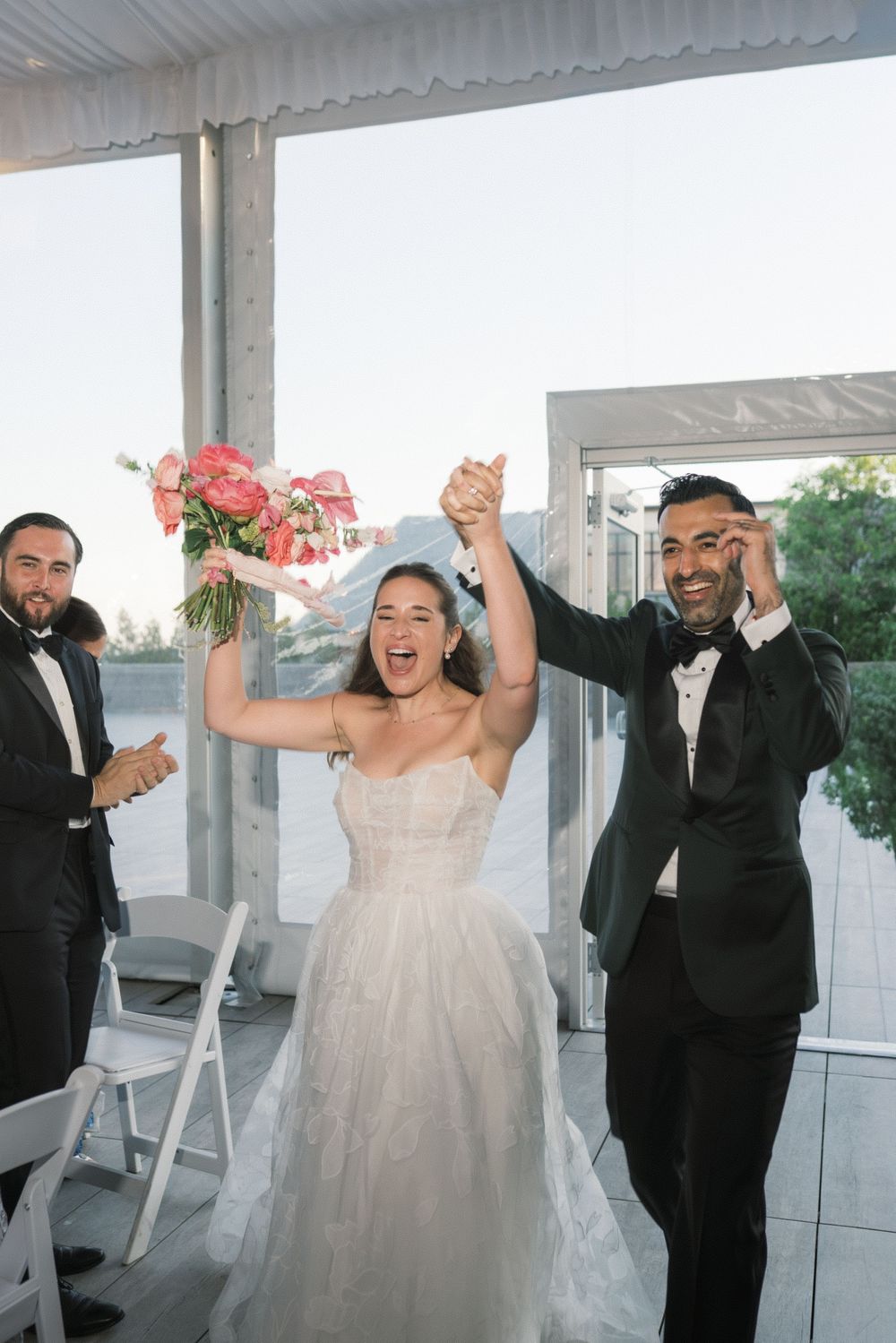 Joyful bride raises bouquet of pink flowers as groom cheers during wedding ceremony exit on rooftop venue.