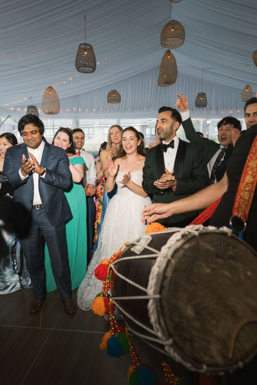 Bride and groom celebrate with guests near a dhol drummer during a festive multicultural wedding reception.