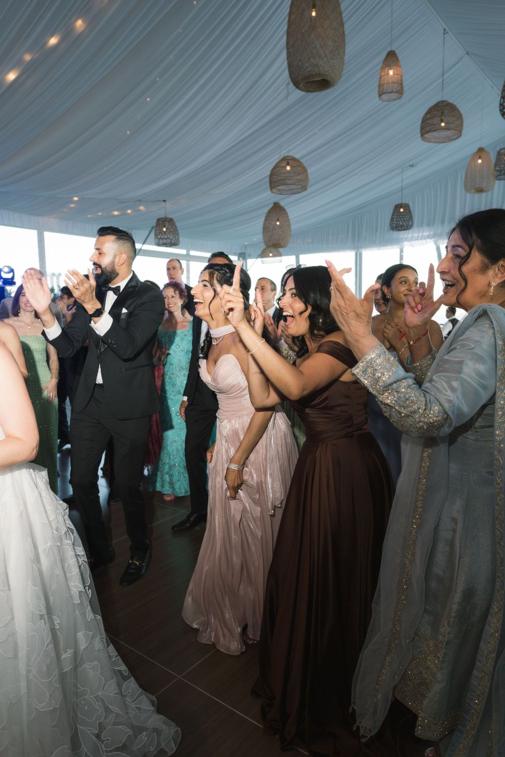 Wedding guests in formal attire celebrate and cheer on a decorated dance floor under hanging lanterns.