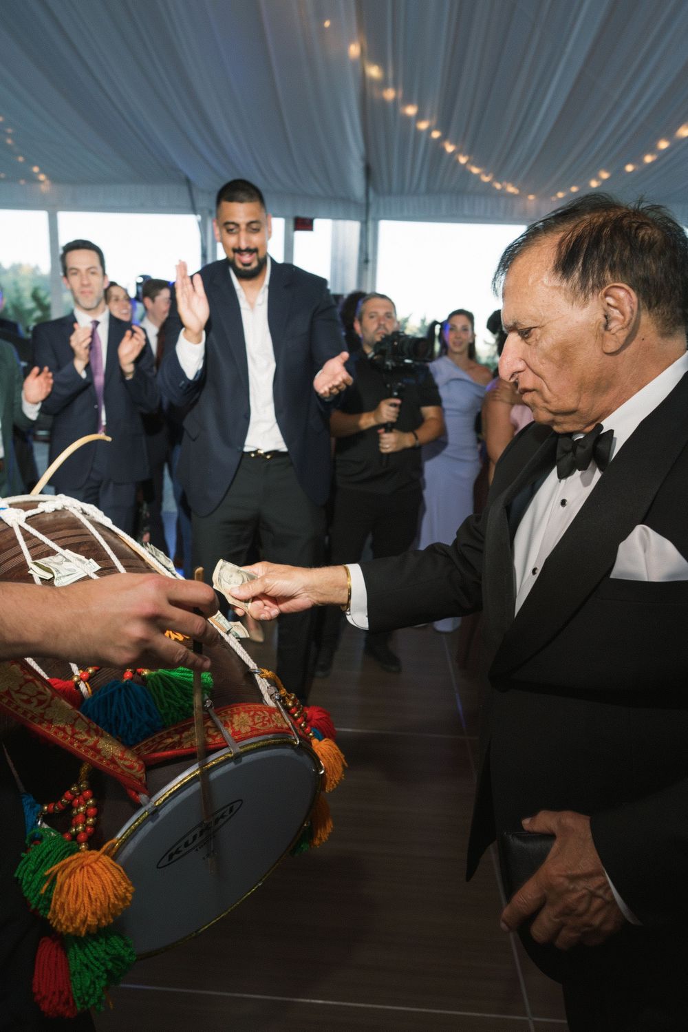 Elderly man in tuxedo tips a colorful dhol drummer at an elegant wedding reception celebration.