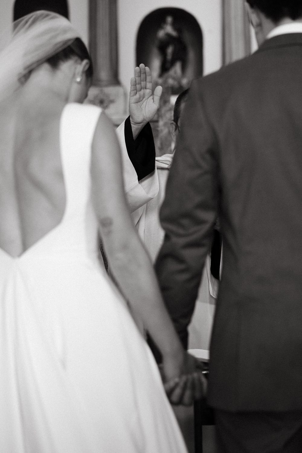 Priest raises hand in blessing over bride and groom holding hands during church wedding ceremony.