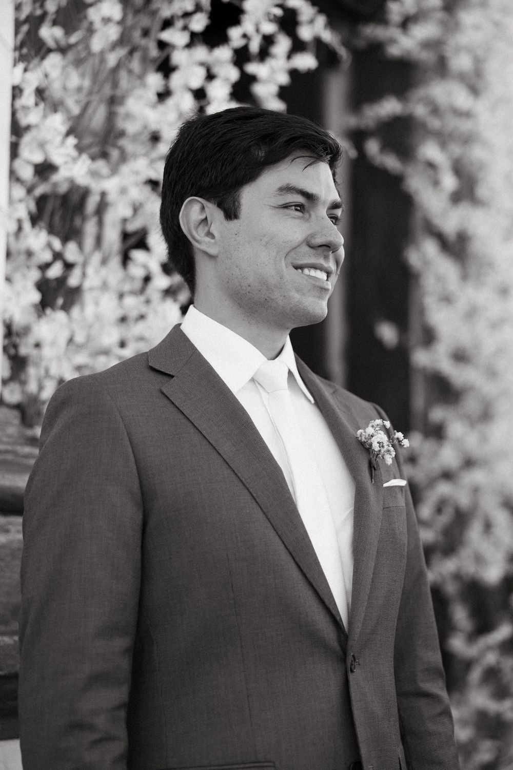 Smiling groom in gray suit with boutonniere standing outdoors among blooming trees on his wedding day.
