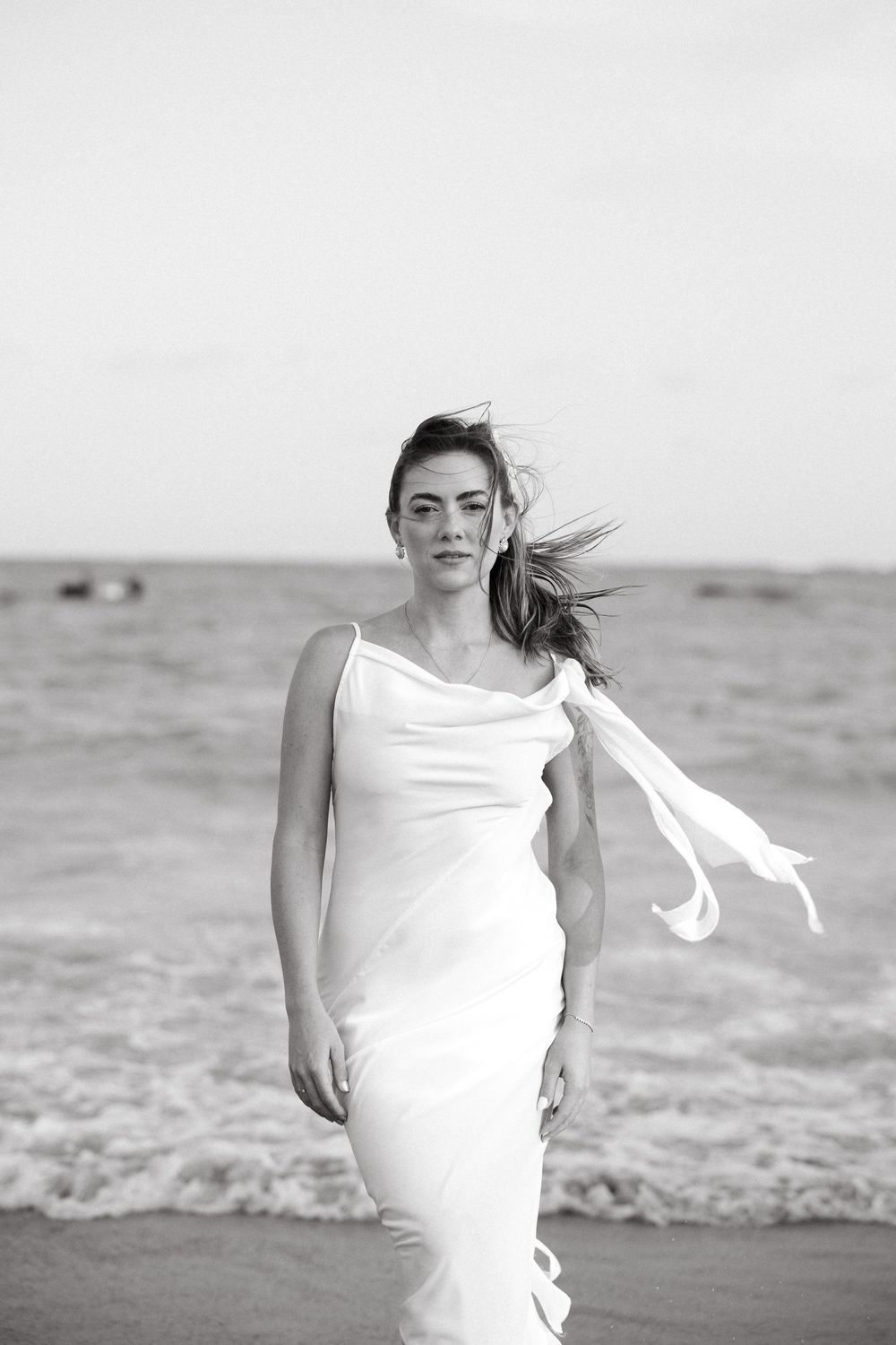 Black and white photo of a bride in a white dress walking barefoot on a windy beach.
