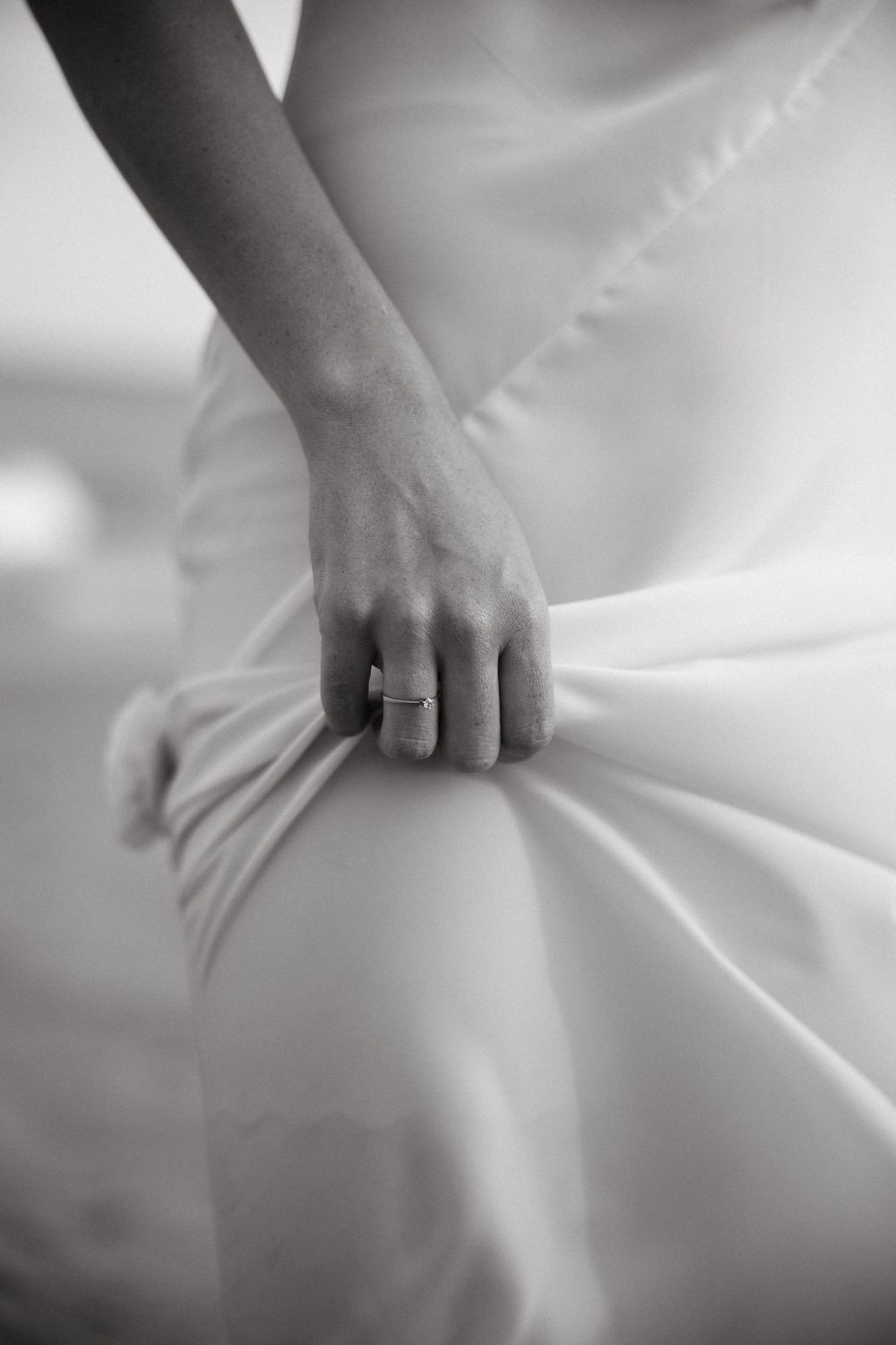 Black and white close-up of a bride's hand holding her white wedding dress, wearing a ring.