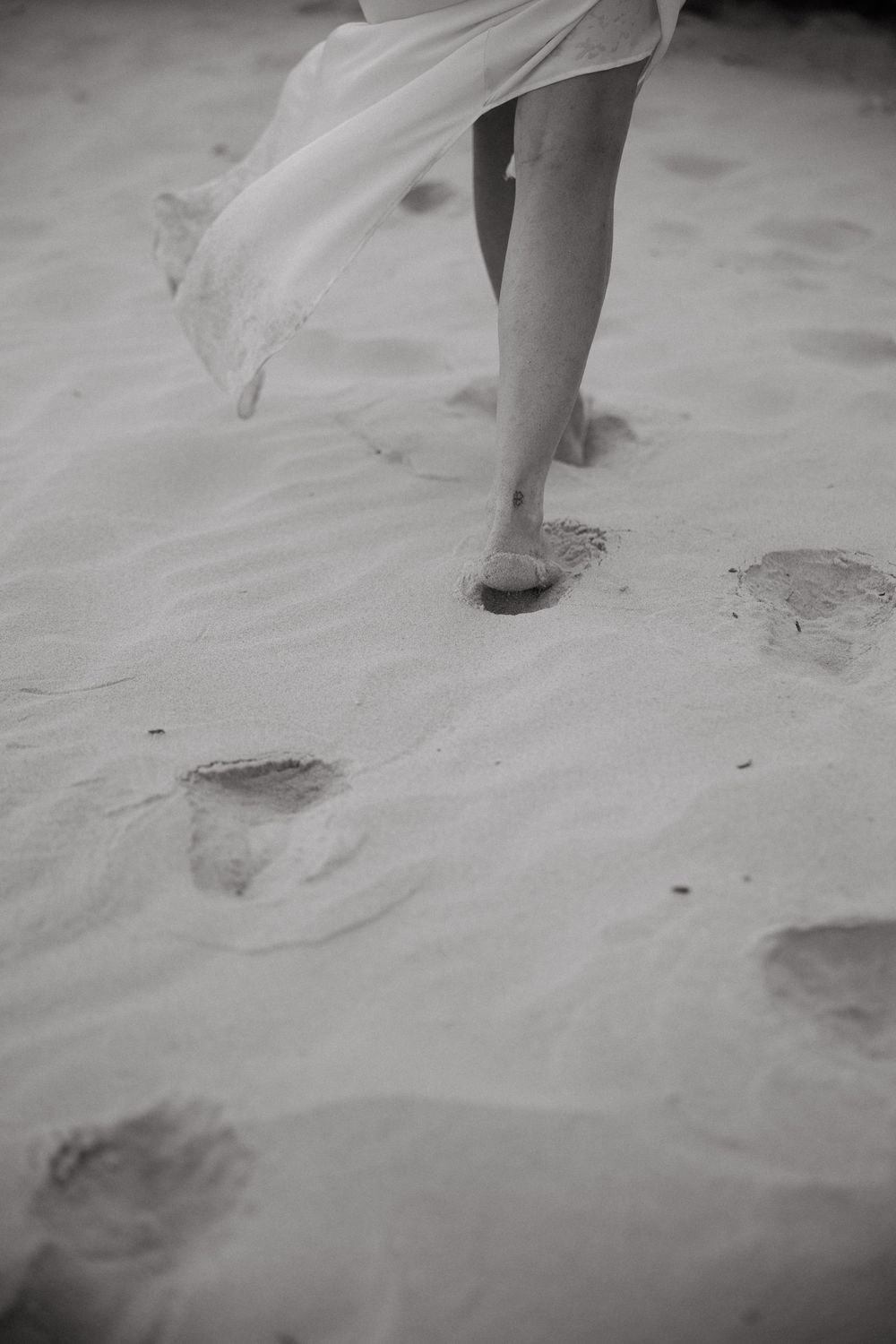 Black and white photo of a bride's bare feet walking on sandy beach leaving footprints.