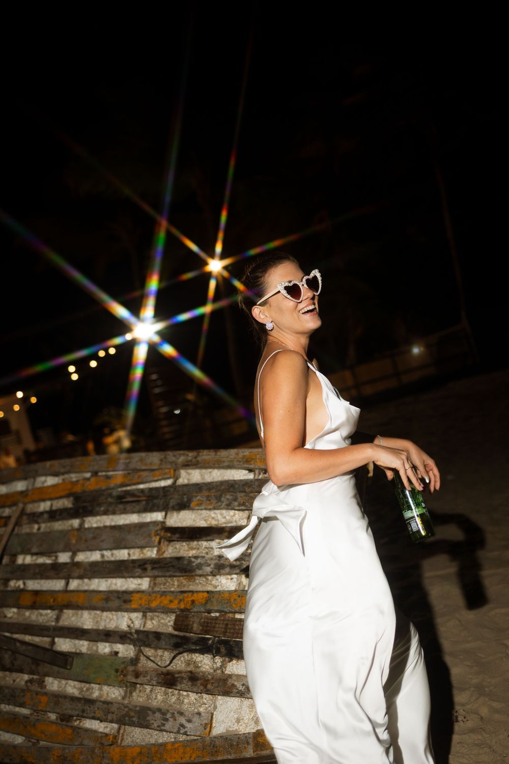 Laughing bride in white dress and heart sunglasses posing outdoors under sparkling night lights.
