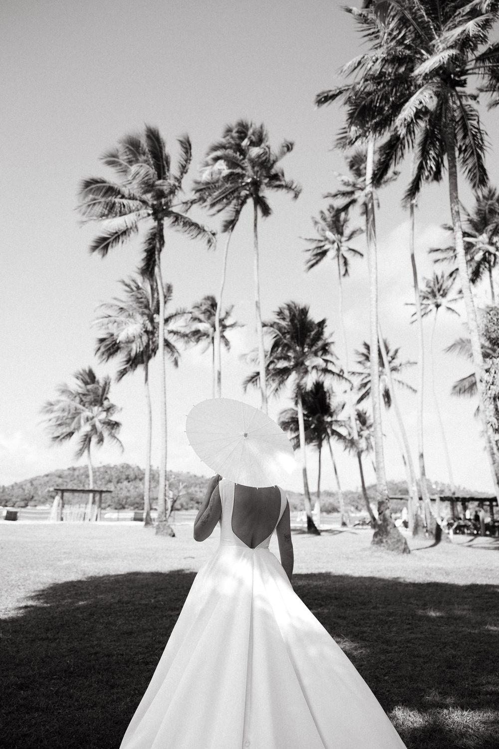 Bride in open-back wedding gown holding white parasol surrounded by tall tropical palm trees.