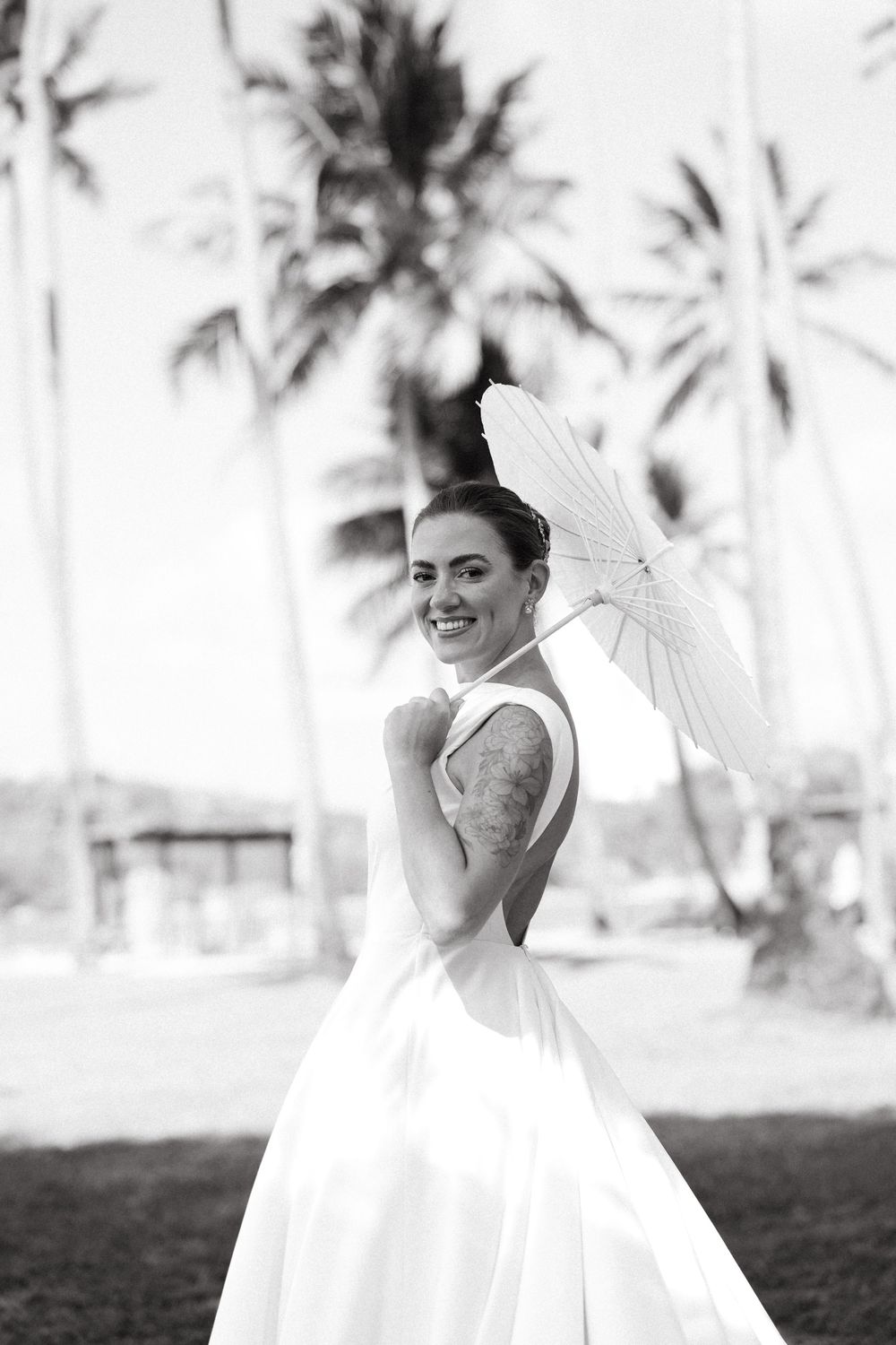 Smiling bride in a white ball gown holding a parasol with tall palm trees in the background.