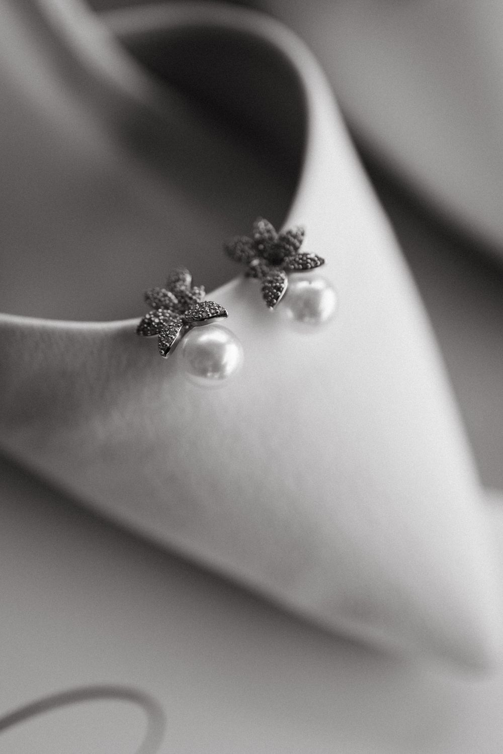 Black and white close-up of elegant pearl and crystal flower earrings resting on a satin bridal heel.