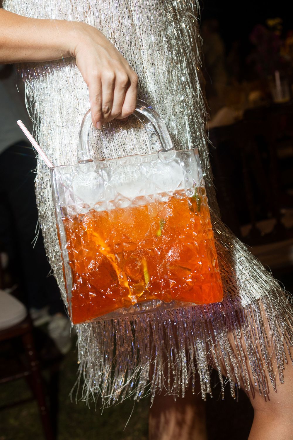 Woman holding a unique transparent handbag filled with orange cocktail drink at a party.